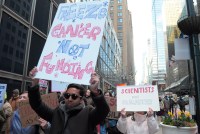 Demonstrators march in a street holding signs, including one that says "Freeze cancer, not funding"