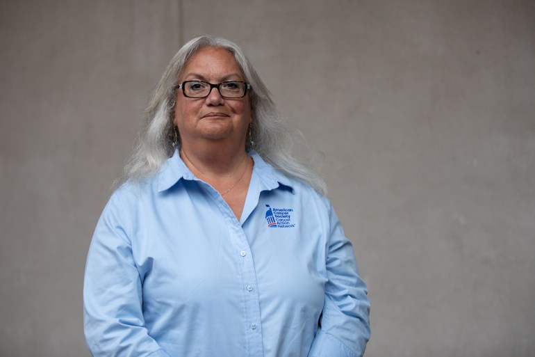 A portrait of an older woman wearing a blue shirt standing against a gray background.