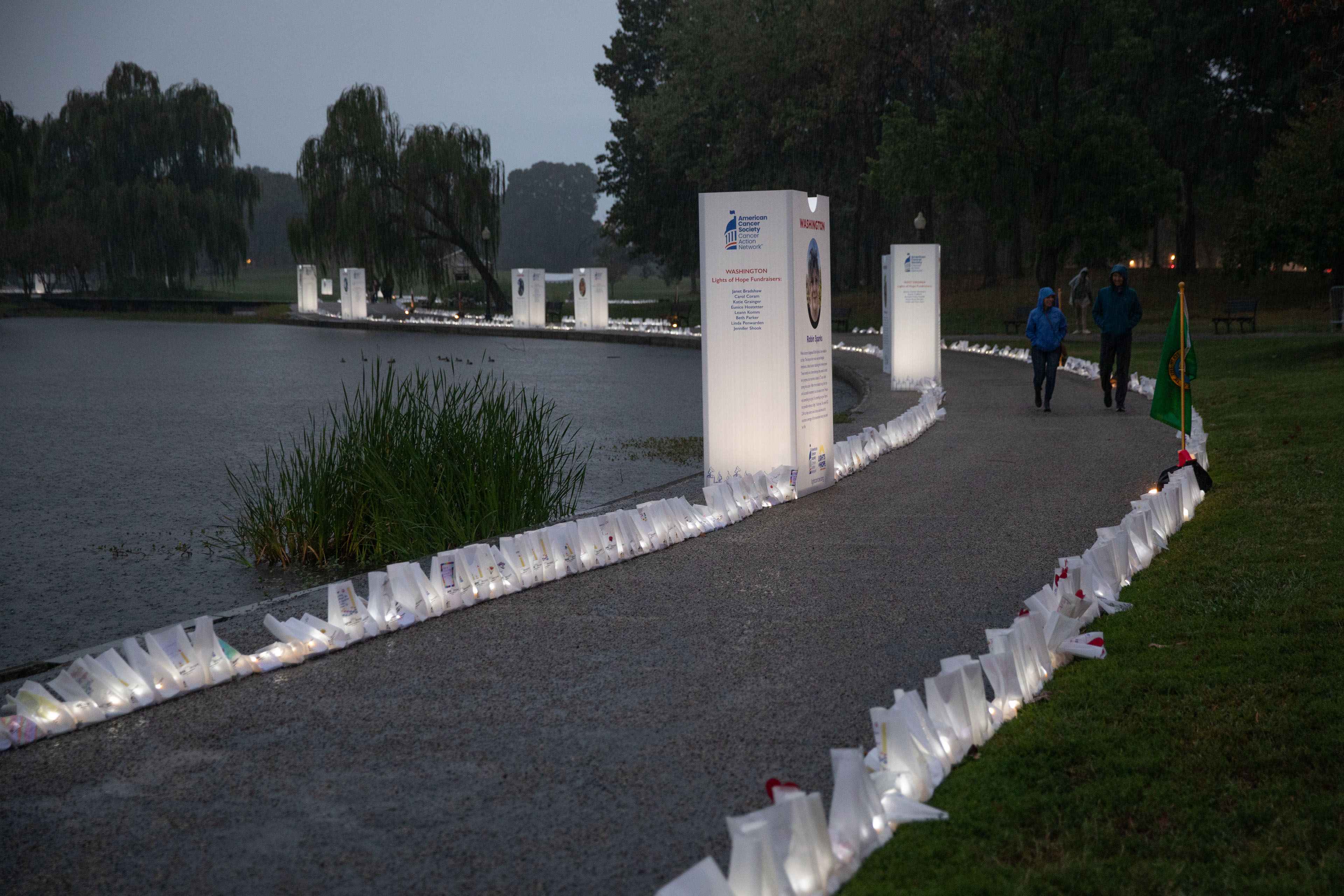A photograph of a path alongside a body of water. The path is lit with white paper bags that are memorials for people who passed from cancer.