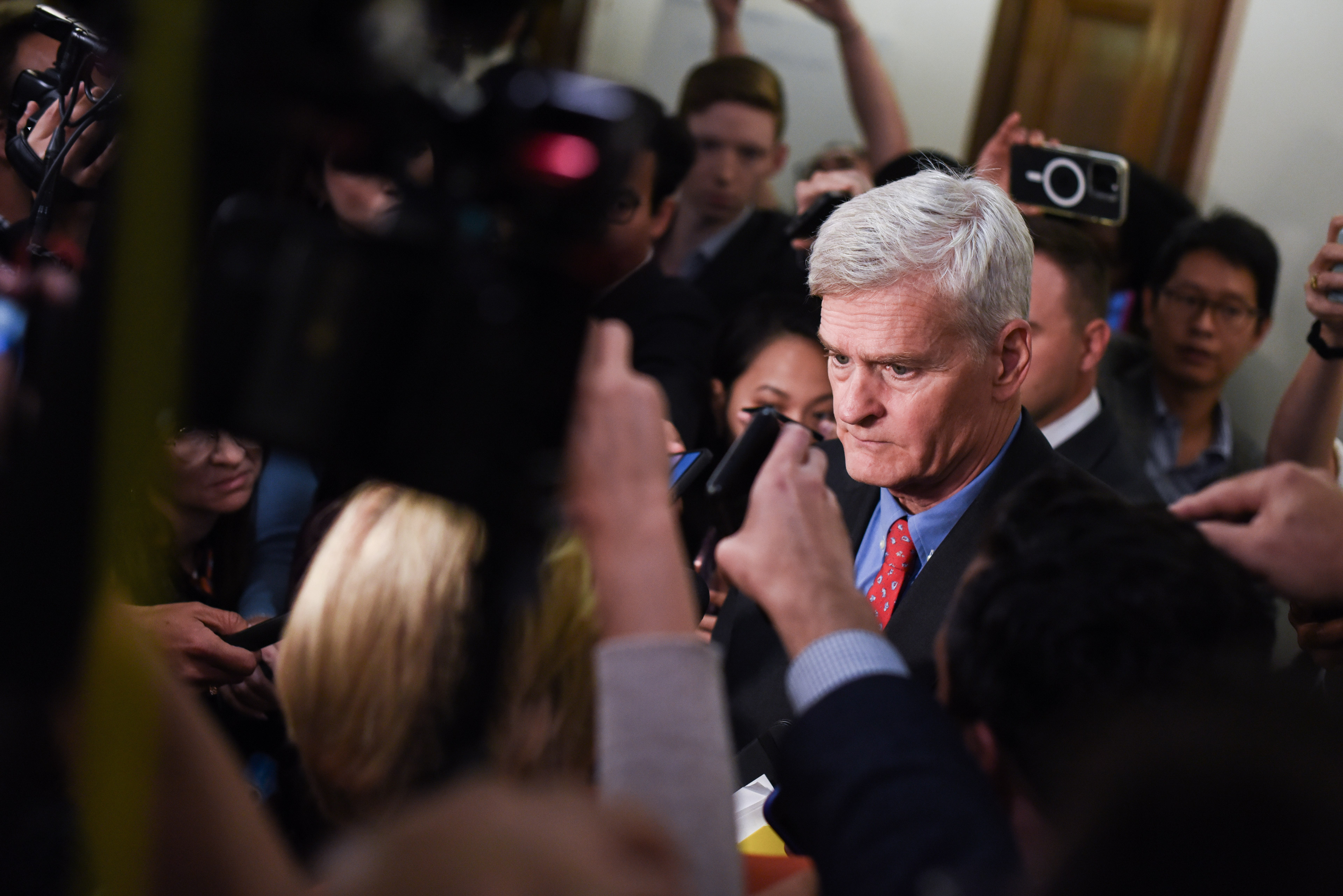 A gaggle of reporters holding up their phones and video cameras interview Senator Bill Cassidy in a Senate hallway.