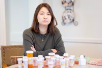 A photo of a woman sitting at a table with several pill bottles in front of her.