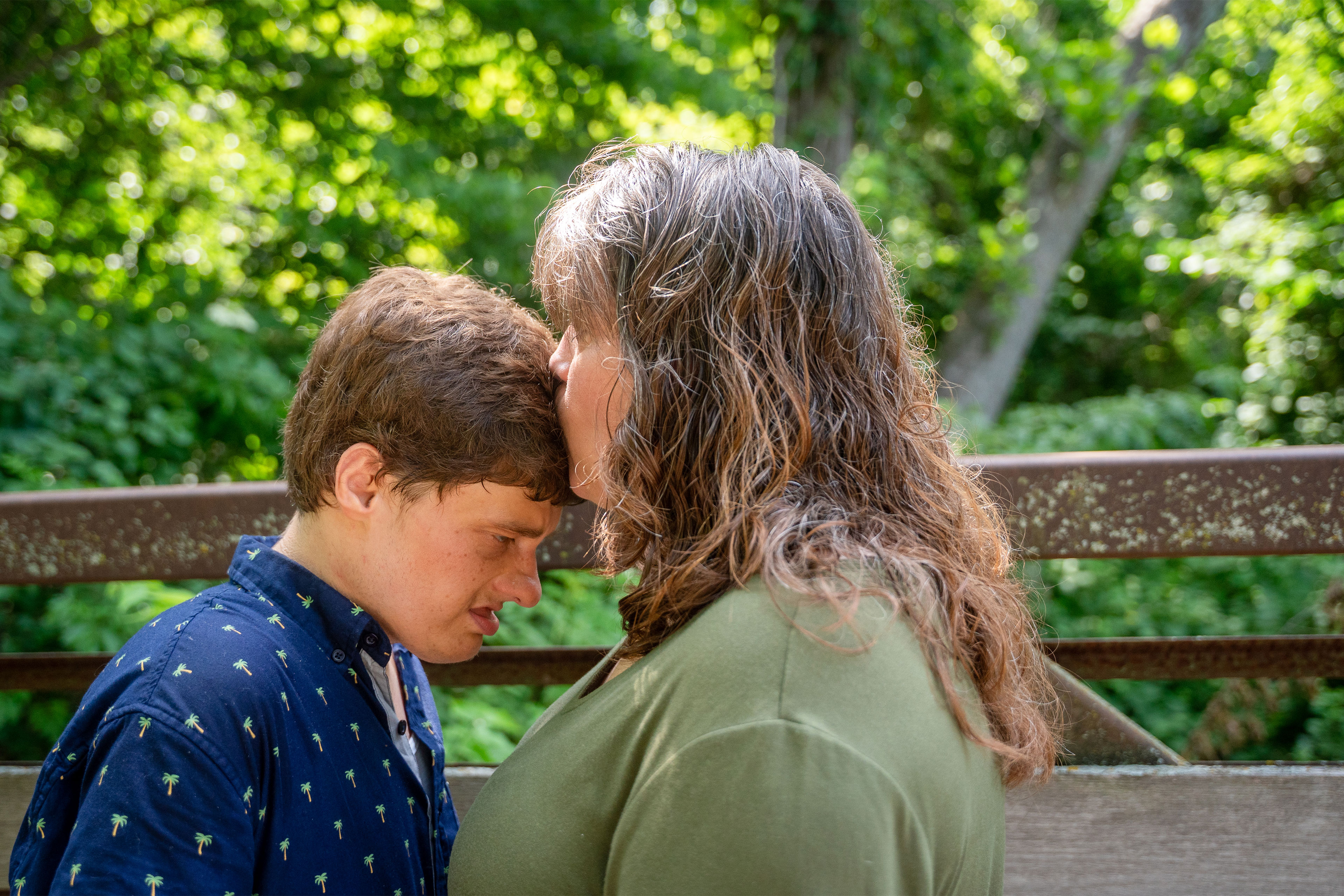 A woman wearing an olive green shirt kisses the forehead of a young man wearing a dark blue shirt with small palm trees on it. The two are standing on a footbridge in a wooded area.