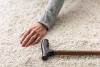 A cropped view of a senior woman's hand on the floor beside a cane on a carpeted floor.