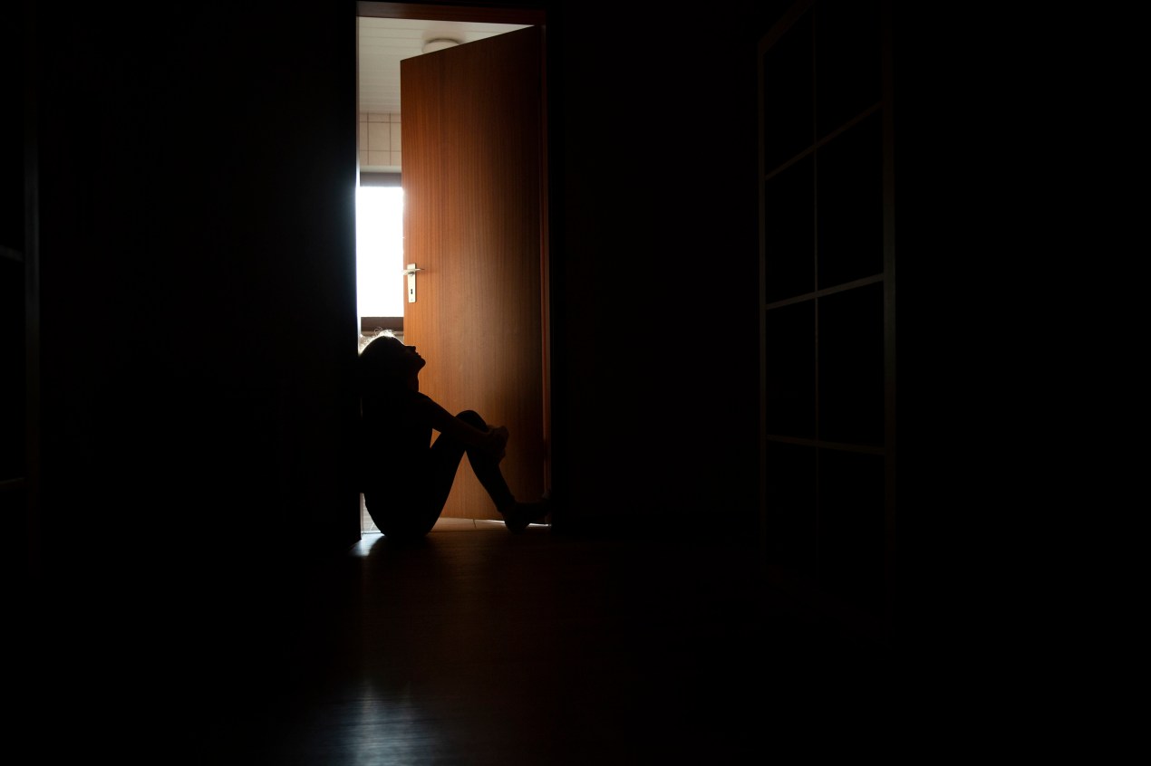 A photo of a teenager sitting in the frame of a doorway backlit by a room filled with daylight.