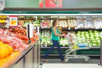 A photo of a woman pushing a grocery cart through a produce aisle in a grocery store.