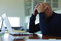 A photo of man holding his head in his hands as he uses a laptop.