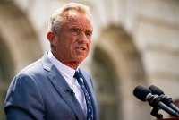 A photo of Robert F. Kennedy Jr. speaking in front of a building. Two microphones are seen next to him.