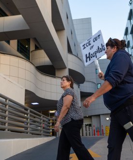 Four people are in frame walking past a sign with white lettering on a red background reading "Emergency Entrance" and "Emergency Department Chest Pain Center" each with arrows pointing right. One person carries a handwritten sign that reads "Keep ICE Out of Hospitals."