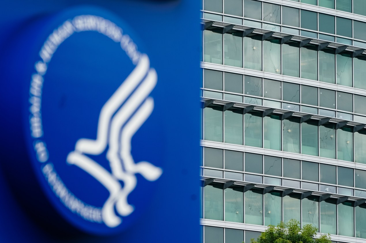 A photo of a large office building. Its windows are riddled with bullet holes. In the foreground is a bright blue sign with an HHS logo.