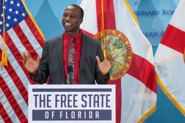 A photo of Joseph Ladapo standing at a podium with the American and Florida flags behind him. A sign on the podium reads "The Free State of Florida."