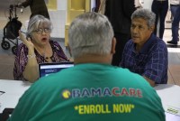 A photo of an older couple seated at a table. A man in the foreground is seen from behind. His shirt reads, "Obamacare. Enroll now."