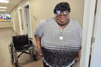 A photo of a woman standing for a portrait in a hallway. A wheelchair is behind her.