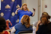 A woman wearing a blue shirt stands in front of a group of people as she gives a presentation