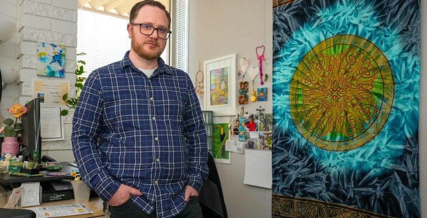 A man wearing a blue plaid button-down shirt and glasses poses for a portrait in an office with a window