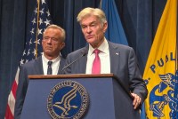 A photo of Mehmet Oz speaking at a podium. Robert F. Kennedy Jr. stands behind him.
