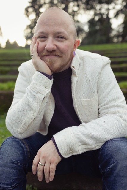 A portrait of a young man wearing a white jacket. he sits with his chin in his palm and smiles towards the camera.
