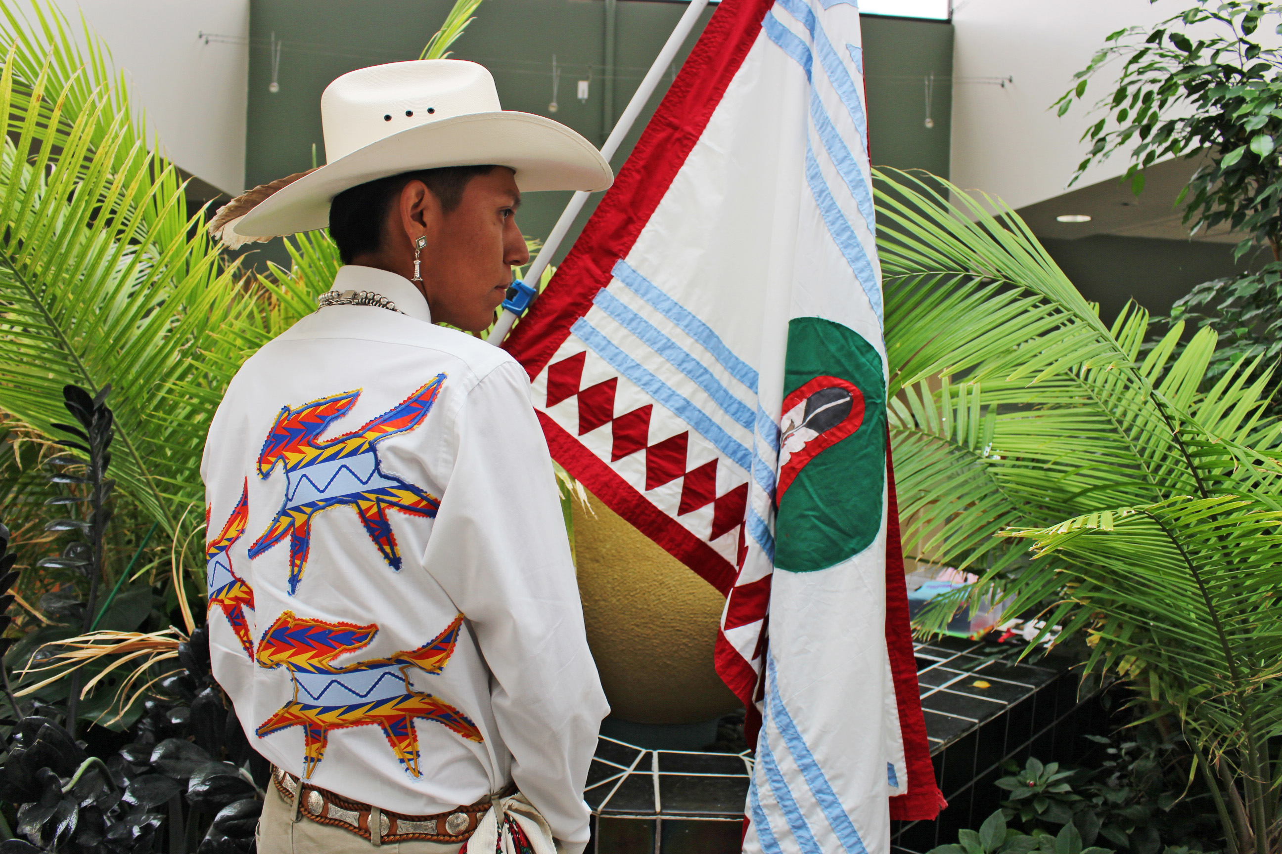 A photo of a man posing for a picture surrounded by ferns. He holds a Western Shoshone flag. The back of his shirt has a custom design of three horses.
