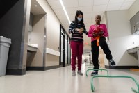A woman wearing glasses and a mask helps another woman balance on one leg while holding a ball
