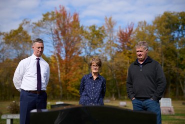 From left, a man, a woman, and another man stand in a cemetery, looking at a gravestone