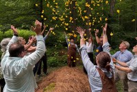 Several people stand around a pile of straw and dirt in a forest, a green burial, and throw yellow petals into the air