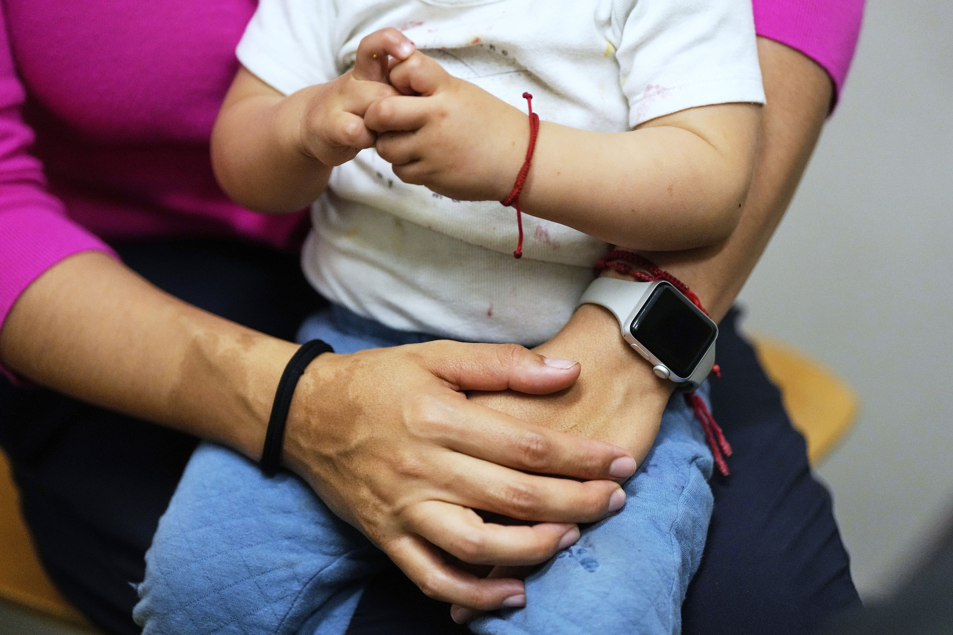 A child wearing a white shirt and a red bracelet sits in the lap of an adult with arms around his waist
