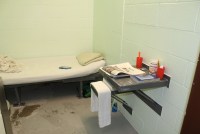 A small prison cell. There is a narrow bed agains the wall with dilapidated sheets, and a metal table connected to the wall.