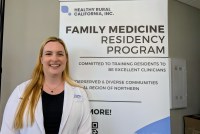 A photo of a female doctor wearing a white coat standing beside a sign that says: "Healthy Rural California, Inc. / Family medicine residency program / committed to training residents to be excellent clinicians."