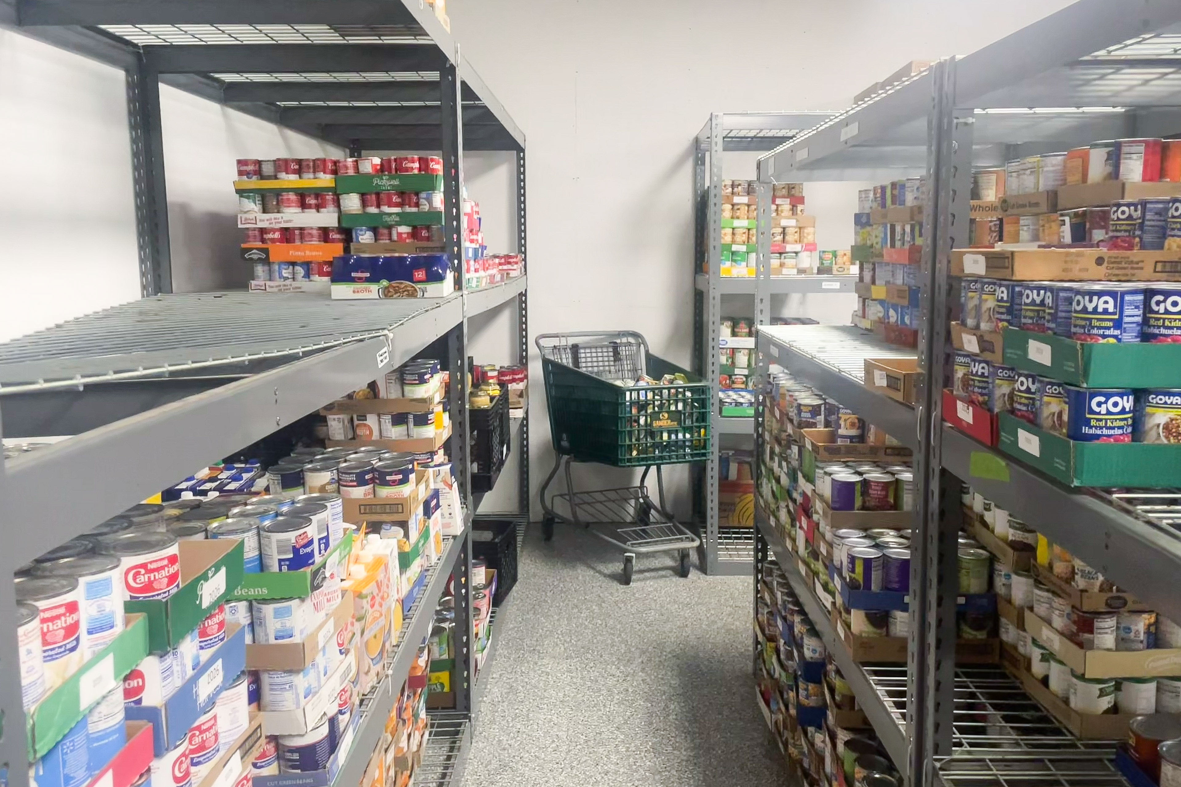A shopping cart inside a food pantry with aisles lined with cans and boxes of goods