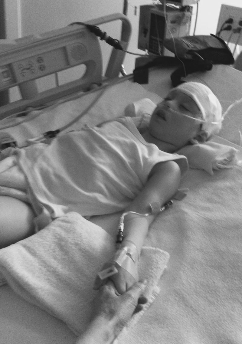 A young boy lays on a hospital bed, an IV in his arm and his head bandaged. The photo is in black and white
