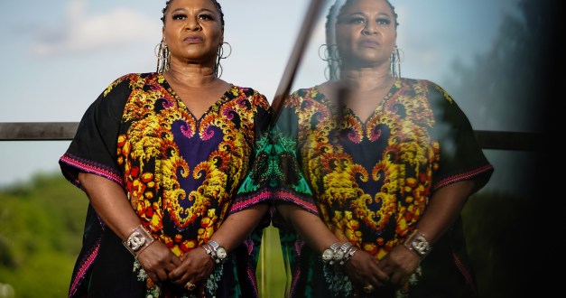 A portrait of a woman leaning against a glass balcony. Her semi-transparent reflection is visible in the right half of the image.