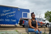 A woman sits in a folding chair in front of a blue van.