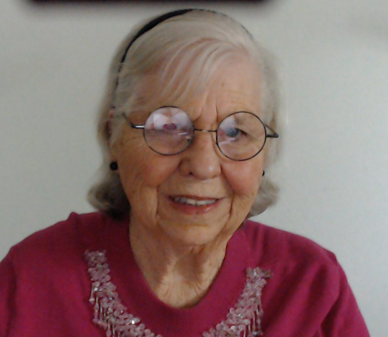 A Zoom photo of a senior woman with short white hair, glasses, and a pink shirt.